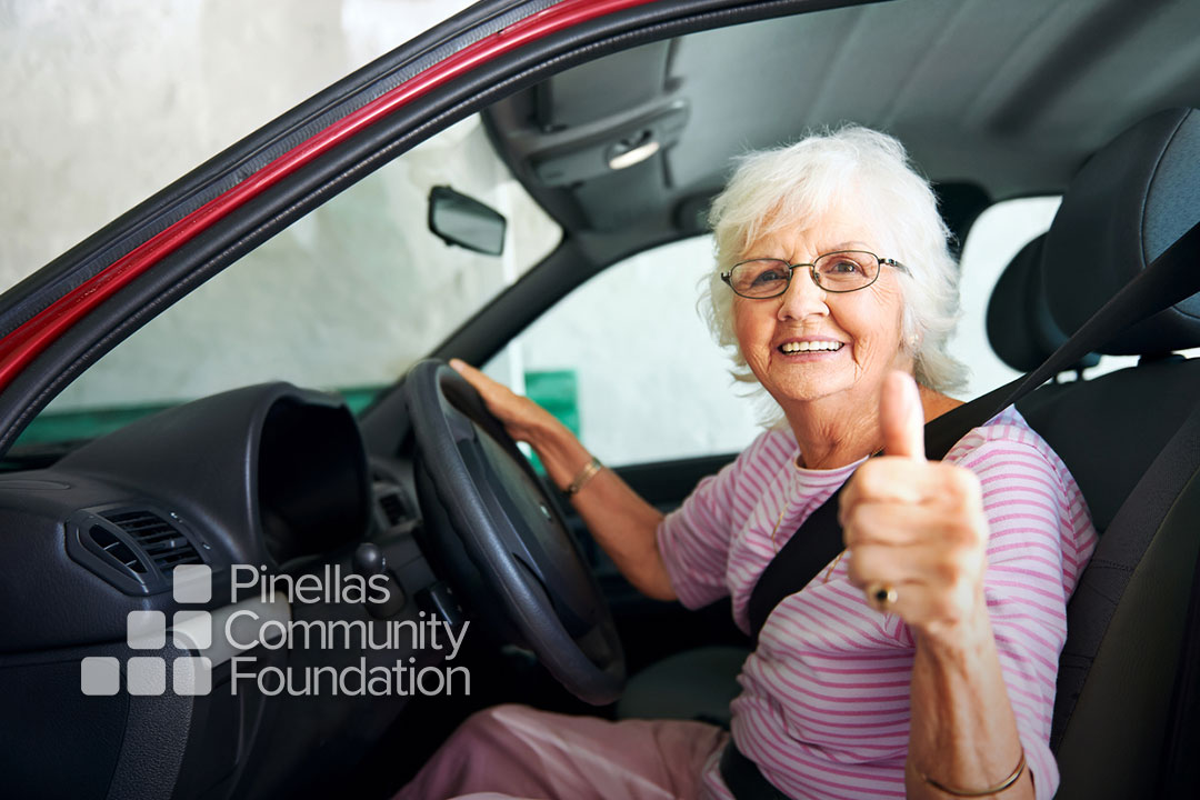 Elderly woman sitting behind the wheel of a car giving a thumbs-up.